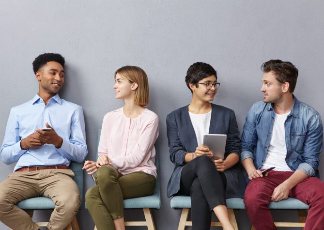 Horizontal portrait of people sit in queue, have pleasant conversation with each other, share ideas and life experience, isolated over grey concrete wall. Diverse group in row, speak and hold gadgets