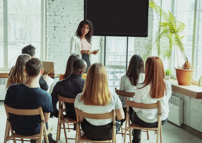 Female african-american speaker giving presentation in hall at workshop. Audience or conference hall. Rear view of participants in audience. Conference event, training. Education, diversity, inclusive concept.