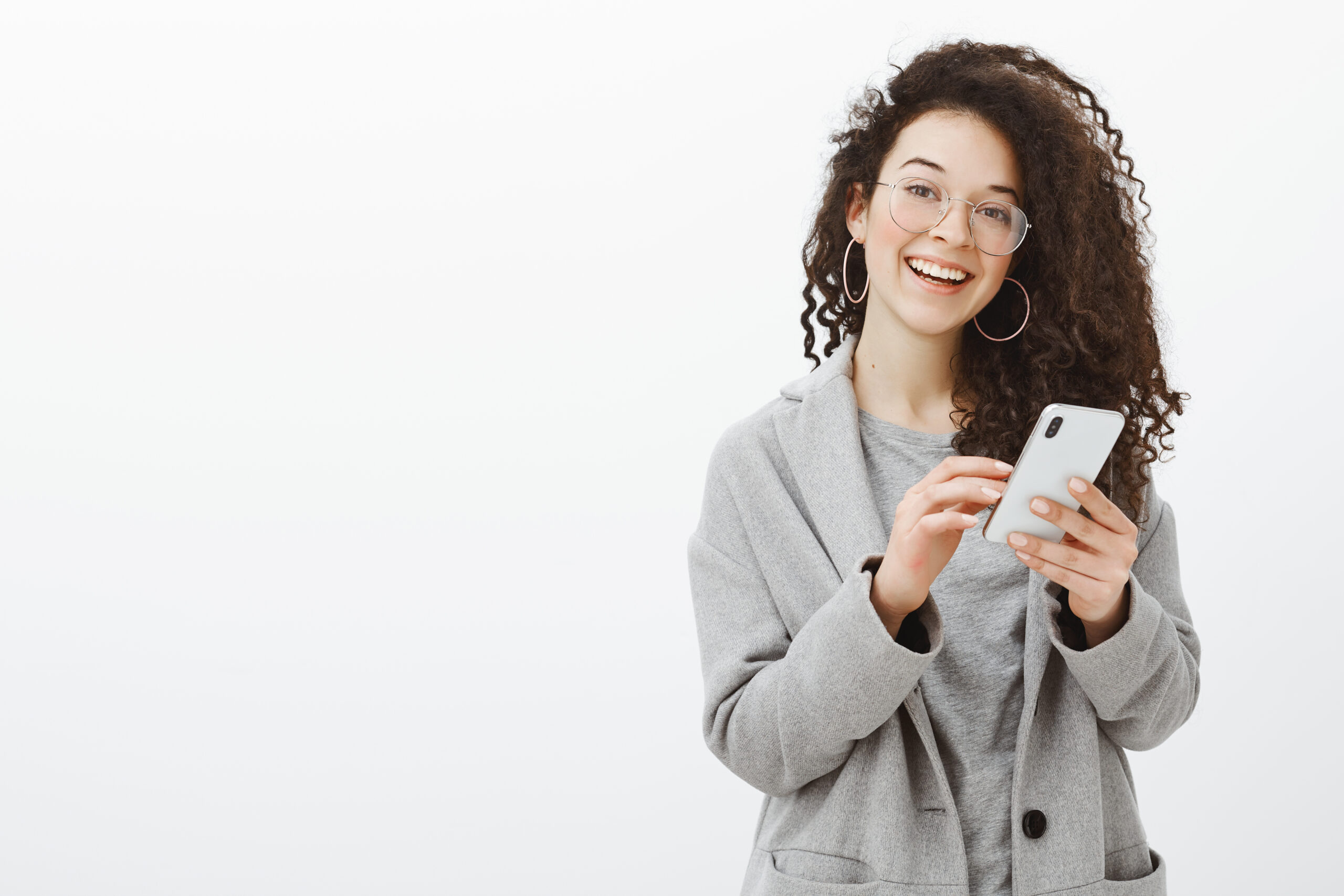 Joyful emotive young european woman in glasses and grey coat with stylish round earrings, holding smartphone and smiling cheerfully at camera while retelling interesting article to friend. Copy space