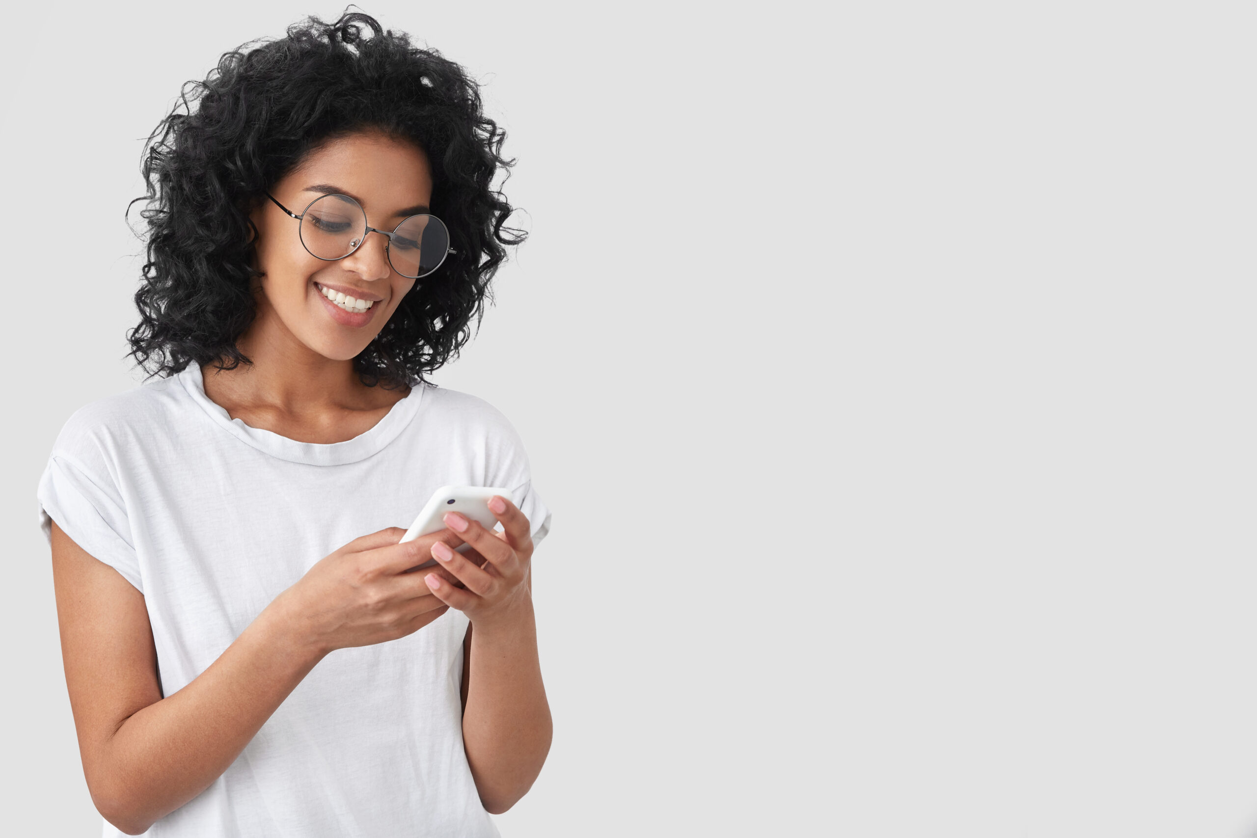 Cheerful African American with crisp hair, holds modern smart phone, happy to recieve message, dressed in casual t shirt, poses against white background with blank space for your advertisement